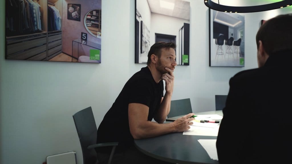 Bearded man in office with LED-lit walls and modern interior posters, sits at a round table with papers, facing an unseen person.