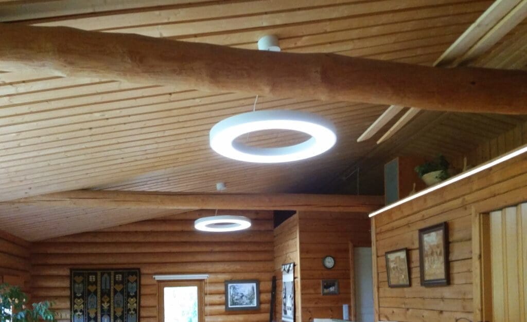 LedStore.fi esittelee modernin LED-valaistusratkaisun rustiikkiin puusisustukseen, jossa on vino katto ja tukevia hirsiä. Kattovalaisimina toimivat kaksi tyylikästä LED-ympyrävalaisinta luovat tunnelmallisen valaistuksen. Seinillä roikkuvat kauniisti kehystetyt kuvat ja elävä tekstiili tuovat lisälämmön tänne kodikkaaseen tilaan.