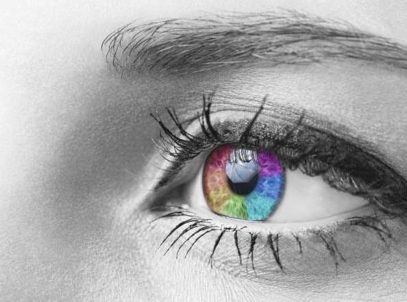 Close-up of an eye with a vibrant, LED-like rainbow iris; black and white background highlights colorful iris. Eyebrow and eyelashes also visible.