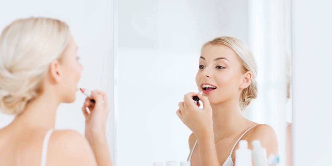 Woman with blonde hair applying lipstick in front of mirror lit by LED lights in bathroom; white top, toiletries visible, bright and soft lighting.