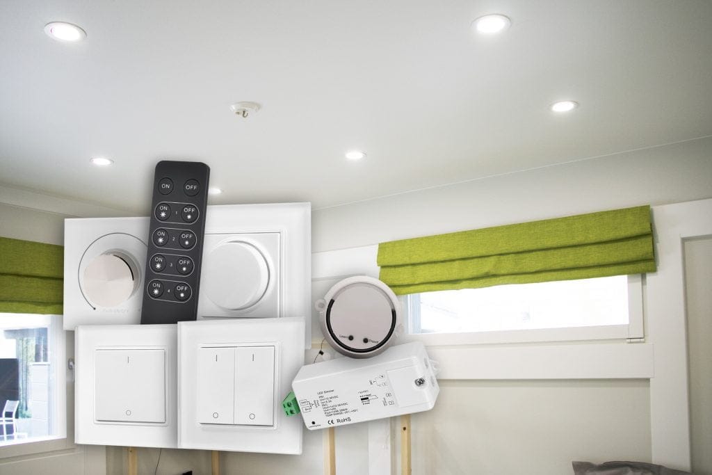 Moderni olohuoneen katto, johon on upotettu LedStore.fi:n LED-valaisimet, valaisee tilaa tyylikkäästi. Alla näkyy kokoelma erilaisia älykotilaitteita, kuten himmenninkytkimiä, kaukosäädin ja relemoduuli. Ikkunaa koristaa vihreä roomalainen kaihdin.