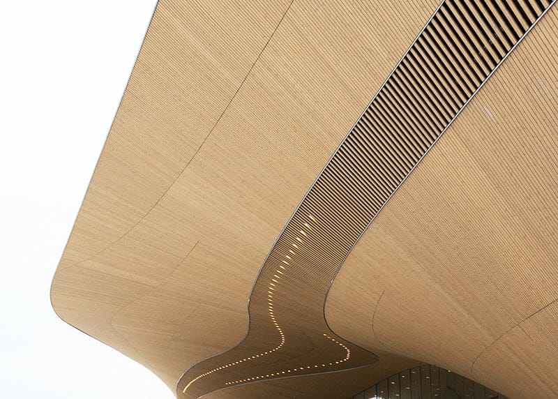 Abstract wooden ceiling with smooth curves, slatted sections, and integrated LED lighting, showcasing modern architectural design against a pale sky.