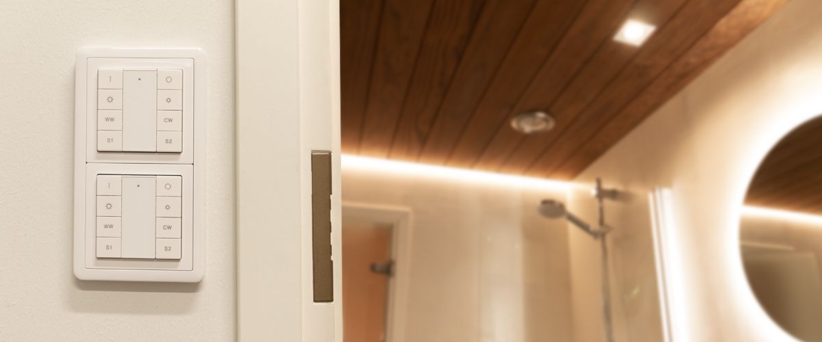LedStore.fi:n valikoimaan sopisi täydellisesti moderni kylpyhuone, jossa on puinen katto ja tyylikäs pyöreä peili sekä suihkualue. Sisäänkäynnin vieressä komeilee LED-seinäpaneeli, jossa on useita valkoisia neliöpainikkeita. Tilassa vallitsee pehmeä ja lämmin valaistus, joka luo miellyttävän ilmapiirin.